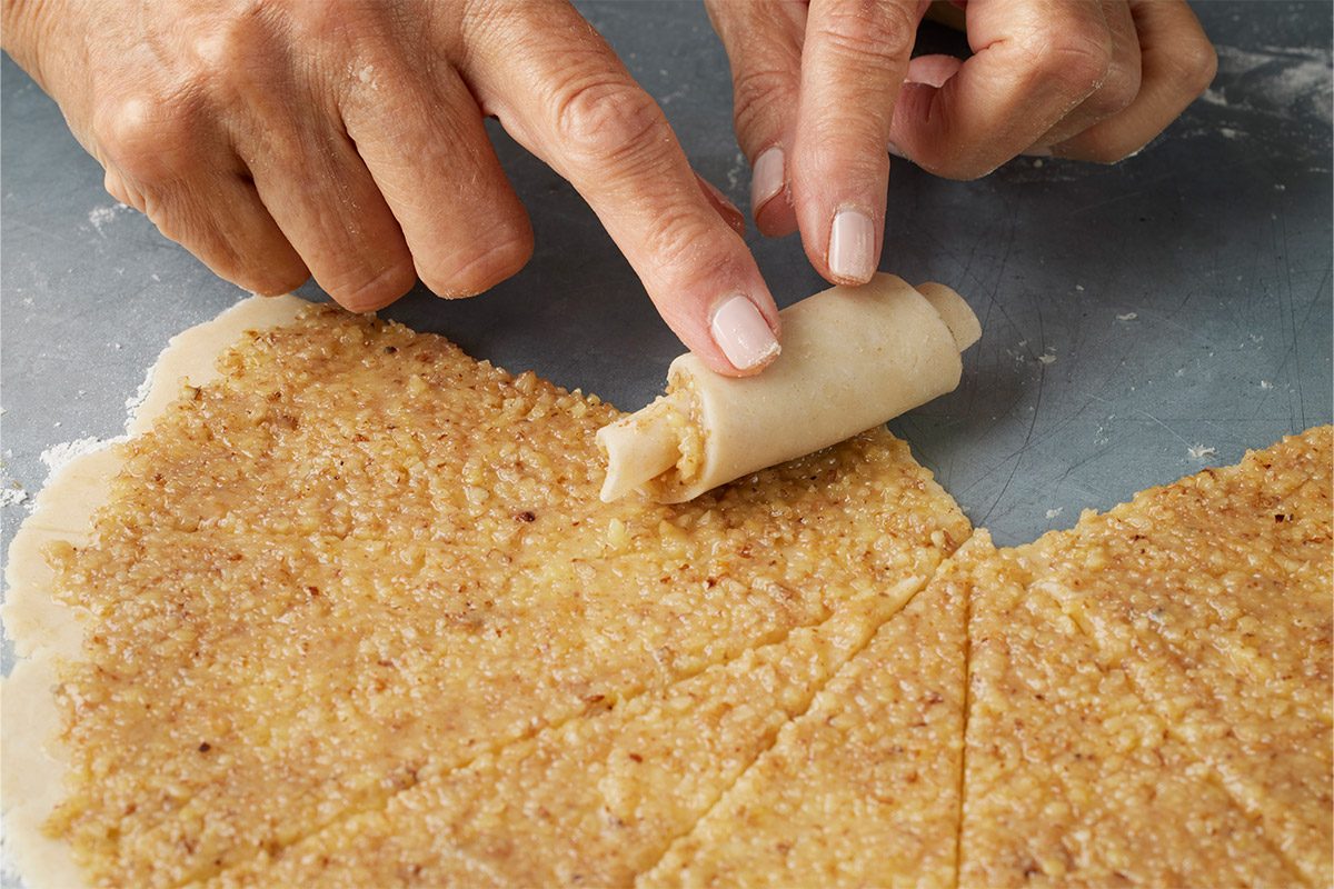 Close-up of hands rolling a triangular piece of dough with a nut filling on a floured surface, preparing pastries;