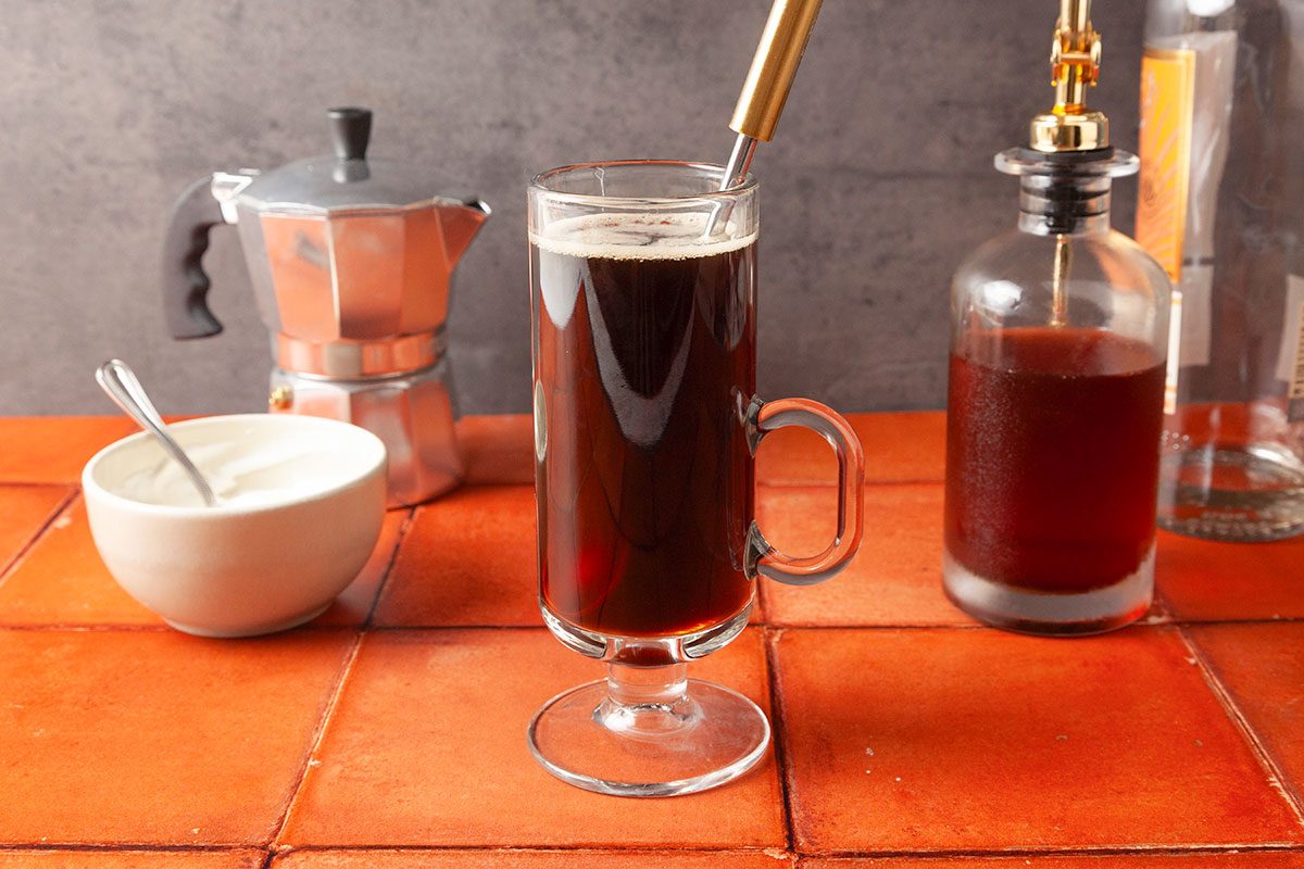 Table view shot of a glass mug filled with black coffee and a metal straw on a tiled counter, surrounded by a moka pot, a bowl with a spoon, and a glass bottle of dark liquid;