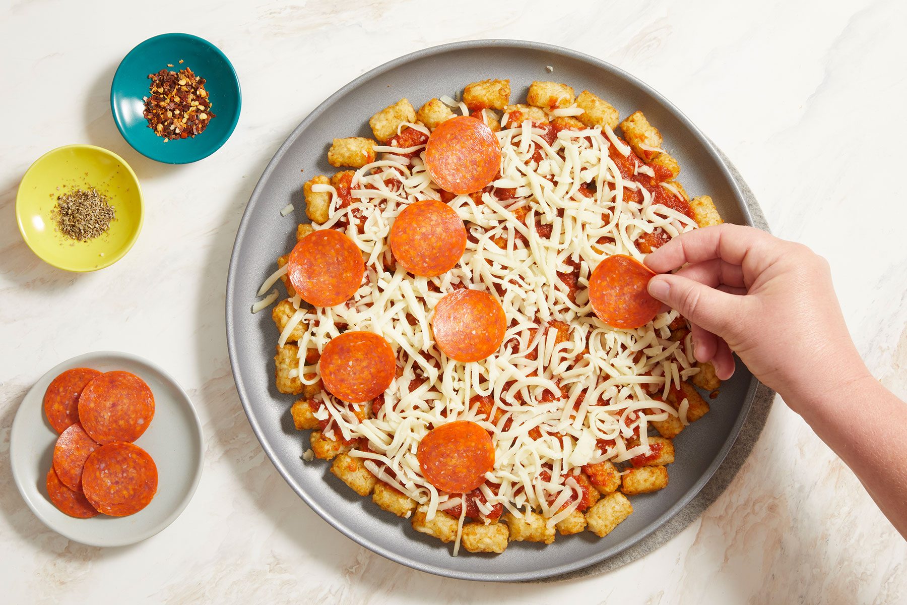 Top the tater tots with cheese and pepperoni in a pan with pizza seasoning and pepper flakes on side.