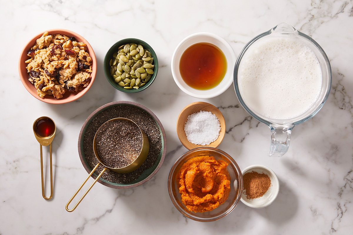 Various ingredients in bowls and measuring cups on a marble surface, including chopped nuts, pumpkin puree, chia seeds, pumpkin seeds, spices, salt, syrup, milk, and vanilla extract.