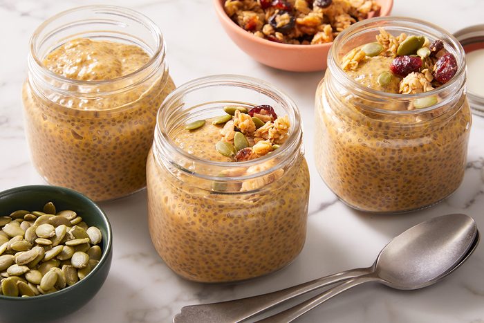 Three glass jars filled with pumpkin chia pudding are topped with granola and seeds, placed on a marble surface. Nearby are a bowl of granola, a bowl of pumpkin seeds, and two metal spoons.