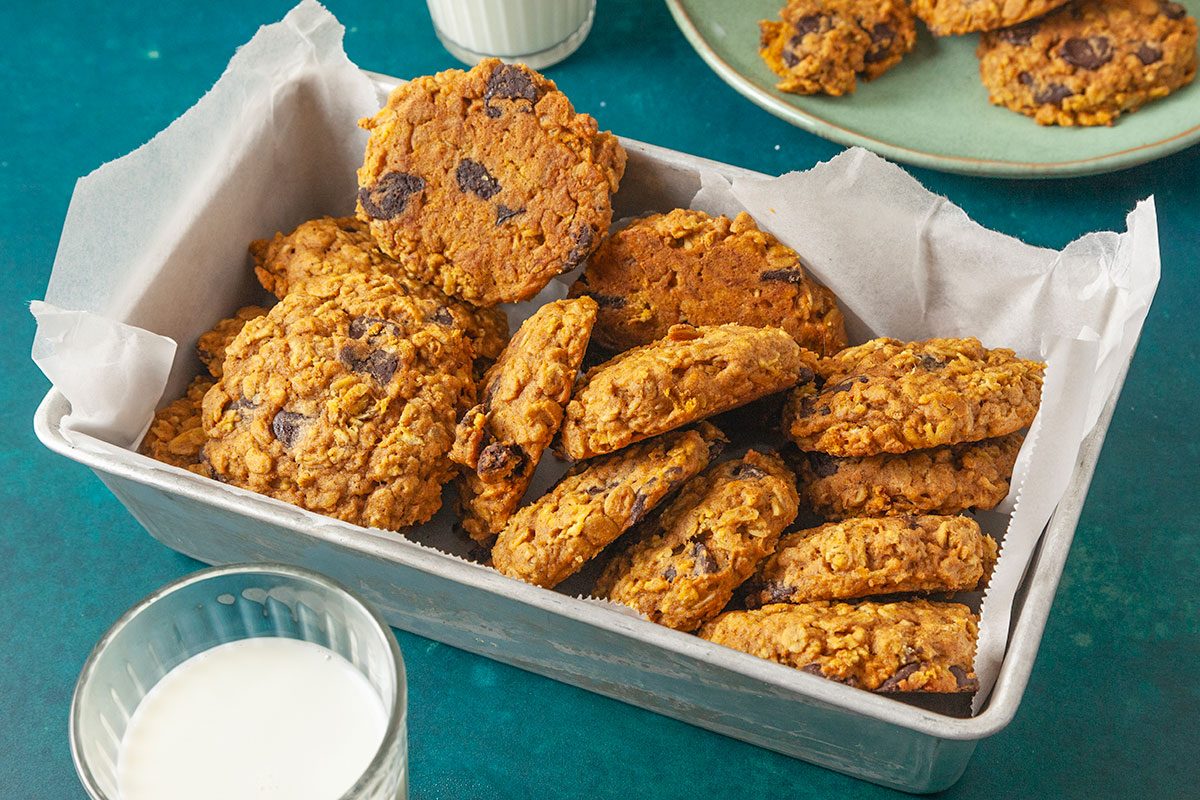 Overhead shot of a metal baking pan lined with parchment paper holding a stack of chunky Pumpkin Oatmeal Cookies; a glass of milk sits nearby, with more cookies on a green plate in the background.