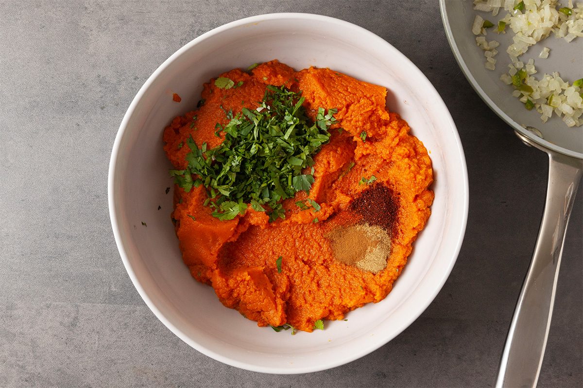 Overhead shot of a white bowl filled with orange mashed sweet potatoes topped with chopped fresh herbs and ground spices, set on a gray surface beside a pan of chopped white onions;