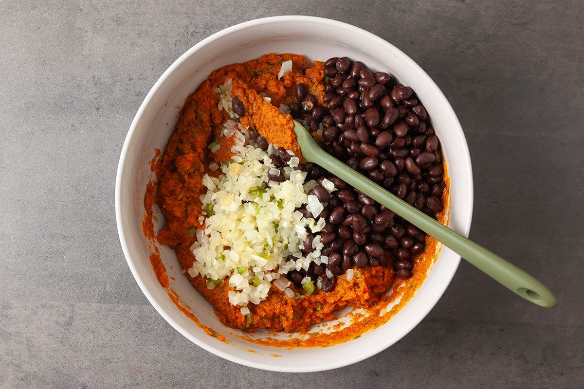 Overhead shot of a white bowl containing mashed sweet potato, black beans, and chopped onions topped with green herbs; a green spoon rests inside the bowl on a gray surface;