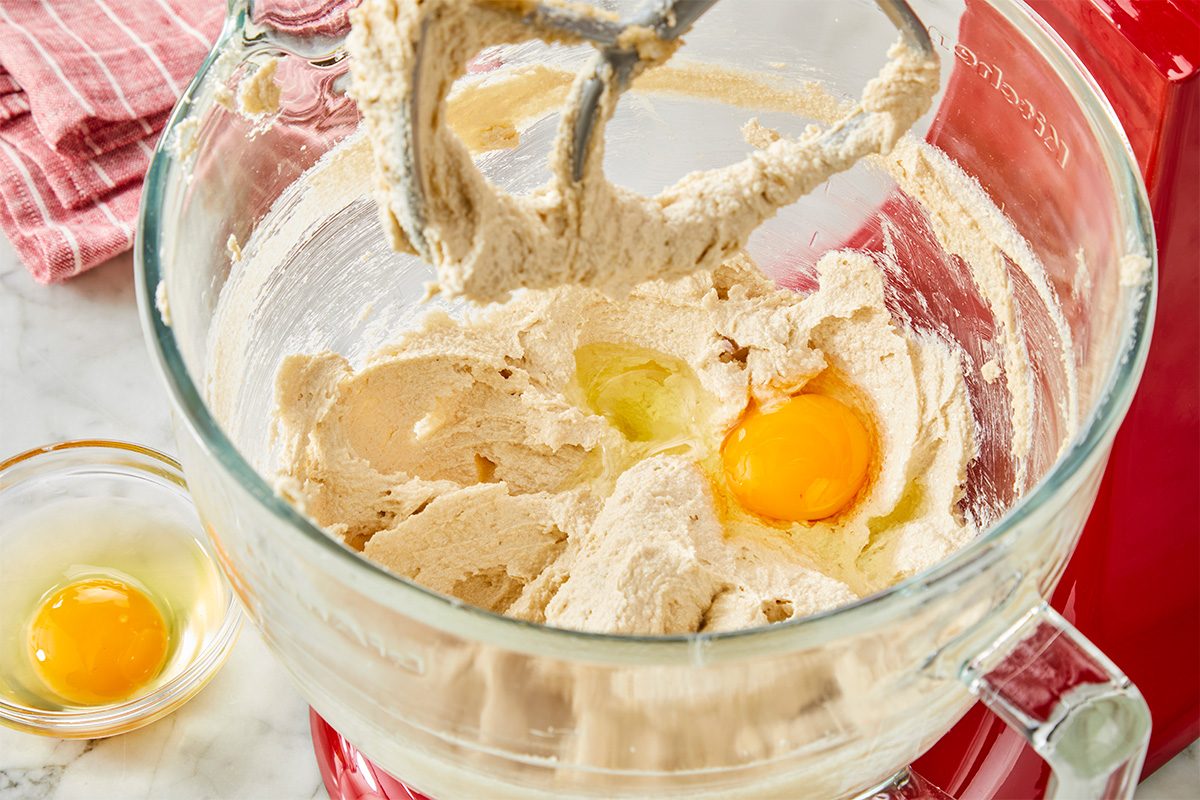 A close-up of a stand mixer with cookie dough, sugar, and butter being mixed. An egg is being added to the bowl, with another cracked egg in a small dish nearby. A red kitchen towel is in the background.