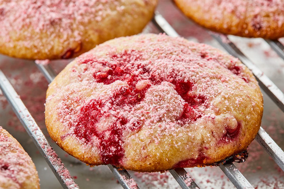 A close-up of a round cookie with visible red berry swirls and a dusting of pink powdered sugar, resting on a metal cooling rack. Other similar cookies are visible in the background.