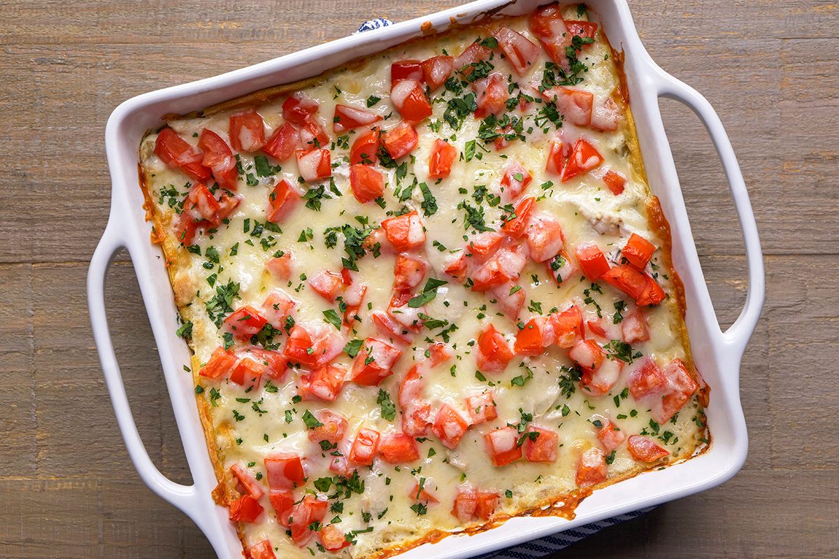 A baked casserole in a white dish, topped with melted cheese, diced tomatoes, and chopped herbs, sits on a wooden surface.