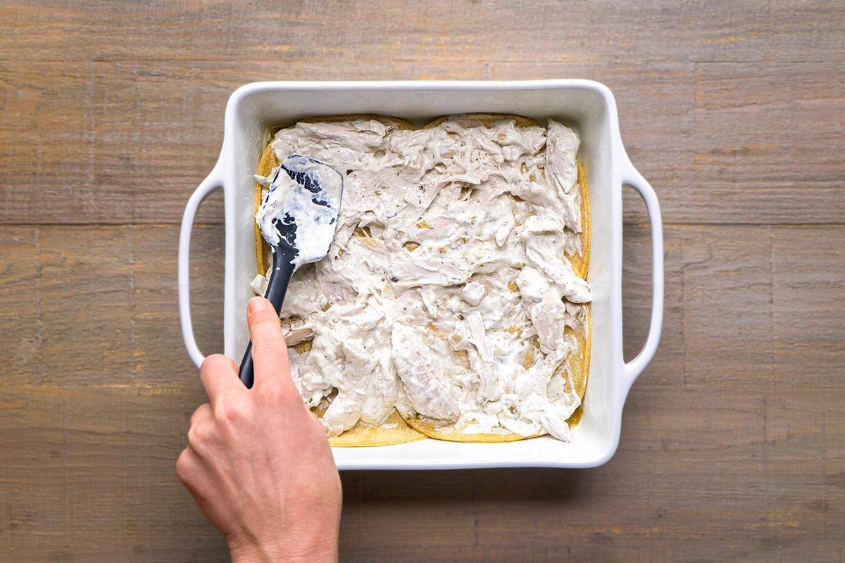 A hand uses a spatula to spread a creamy white mixture over shredded chicken in a square white baking dish on a wooden surface.