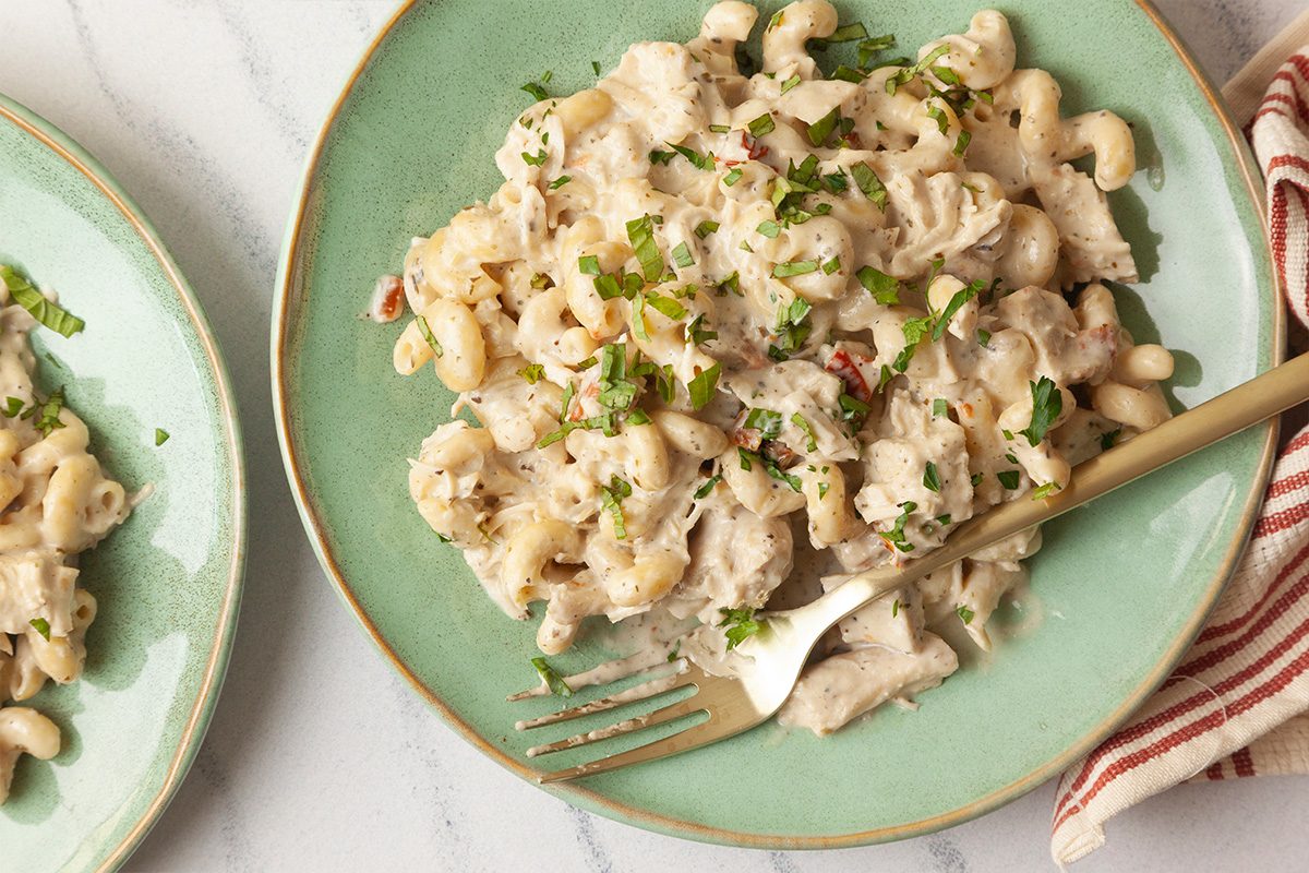 Overhead shot of a green plate of creamy Slow-Cooker Chicken Boursin Pasta mixed with chicken and garnished with chopped herbs, with a fork on the side and a striped napkin nearby