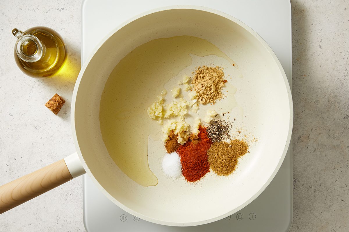 A white pan on a stovetop with olive oil, minced garlic, and various spices including ground ginger, cumin, paprika, black pepper, and salt. A glass bottle of olive oil sits nearby on the countertop.