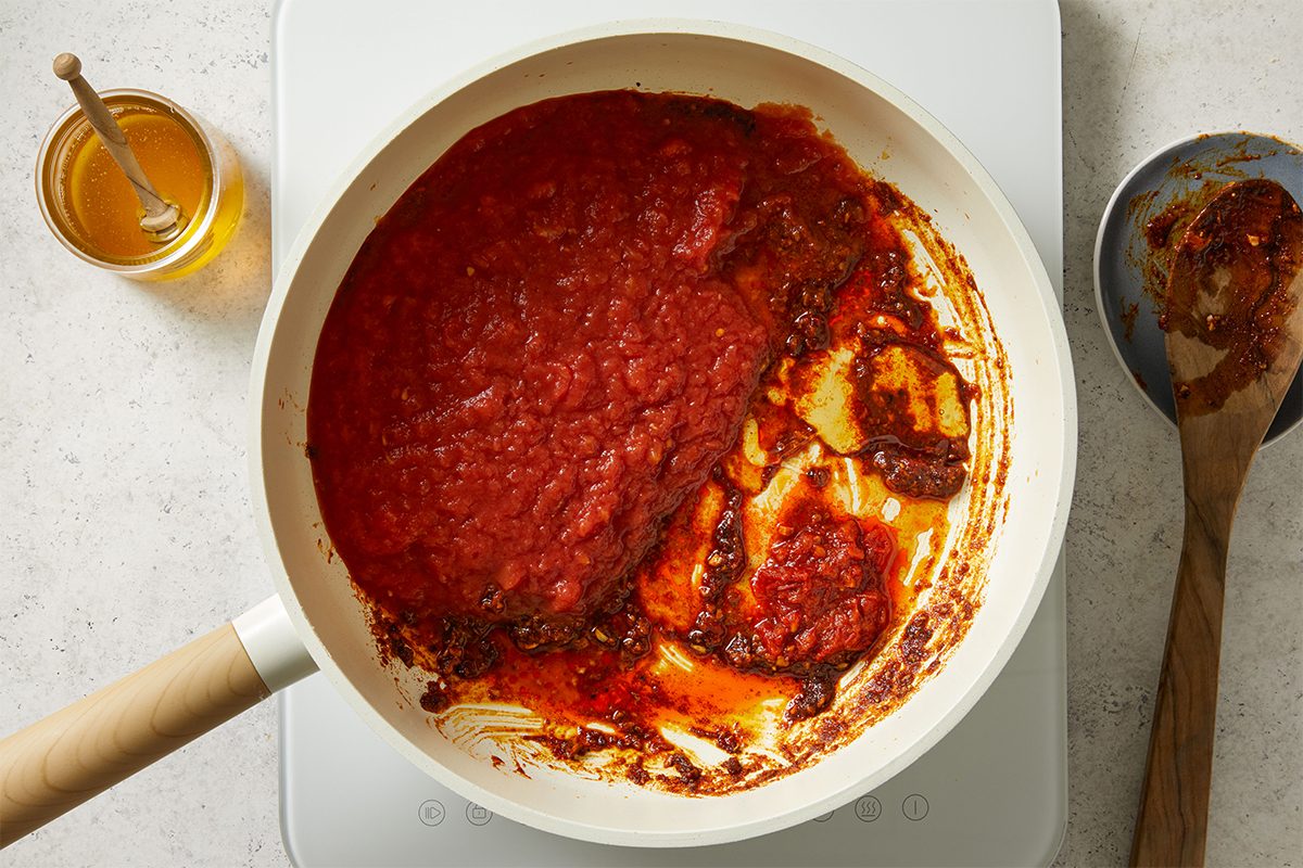 A white skillet on a stovetop contains tomato sauce, with some sauce pushed aside to reveal a caramelized, dark red layer underneath. A wooden spoon with sauce is next to the pan, and a jar of honey is nearby.