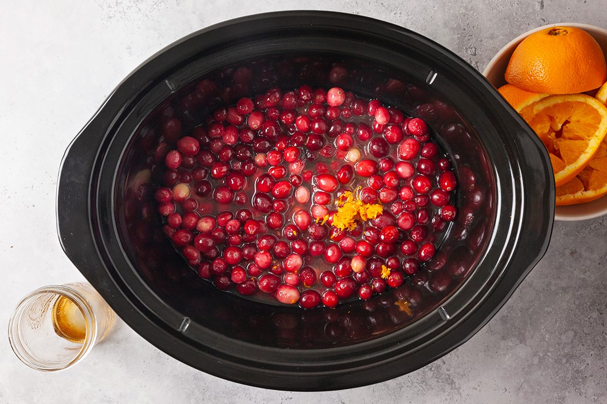 A slow cooker filled with cranberries and orange zest sits on a gray surface next to a bowl of sliced oranges and an empty glass.