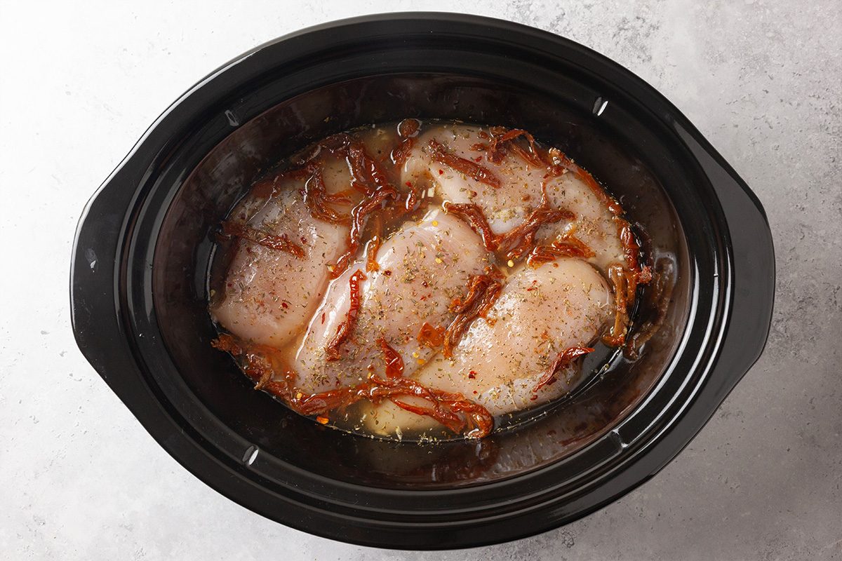 Raw chicken breasts topped with sliced onions, seasonings, and broth are placed inside a black slow cooker, ready to be cooked. The slow cooker sits on a light-colored countertop.