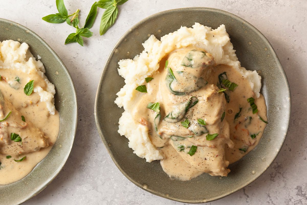 A plate of creamy chicken with herbs served over mashed potatoes, garnished with fresh basil. Another plate with part of the same meal and basil leaves are visible nearby on a light-colored surface.