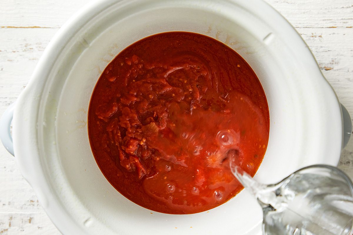 Overhead shot of tomato sauce being poured into a slow cooker with a ladle resting inside.