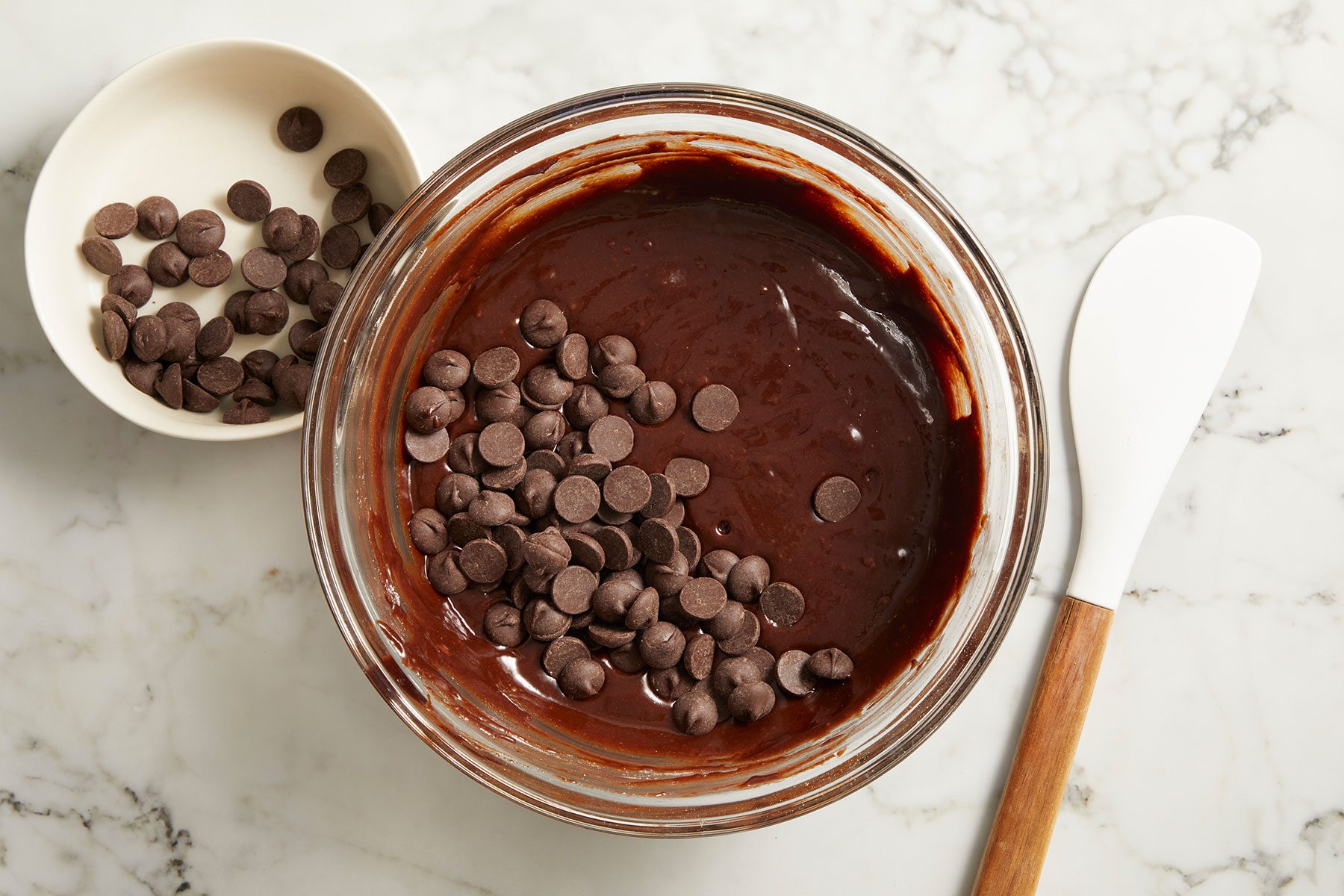 Folding dark chocolate chips in the brownie batter in a large glass bowl with a spatula kept on side.