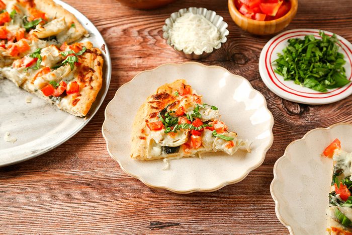 A slice of vegetable pizza sits on a beige plate, surrounded by a whole pizza, chopped green herbs, diced tomatoes, and shredded cheese in small bowls on a wooden table.