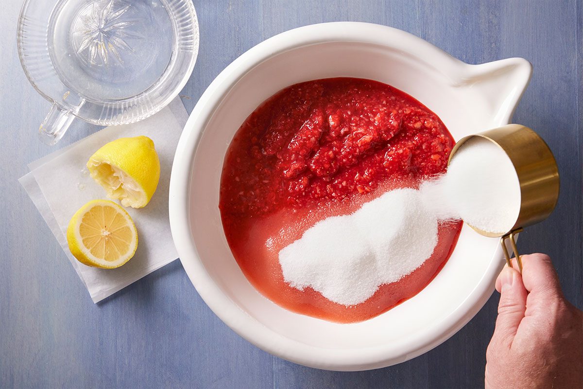 A hand pours sugar from a measuring cup into a bowl of crushed strawberries. Next to the bowl, there is a citrus juicer and a cut lemon on a napkin, all on a blue surface.