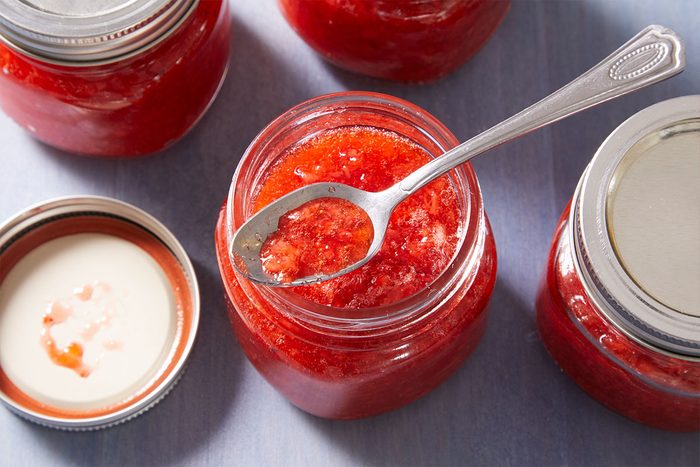 An open jar of bright red jam with a spoon inside sits among other closed jars of jam on a light surface. The lid of the open jar is resting nearby.