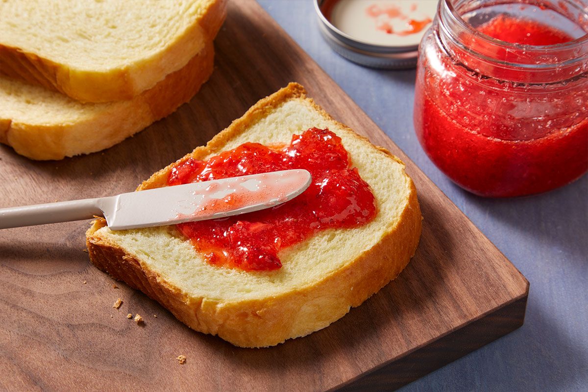 A knife spreads bright red jam on a slice of white bread, with a jar of jam and another slice of bread nearby on a wooden cutting board.