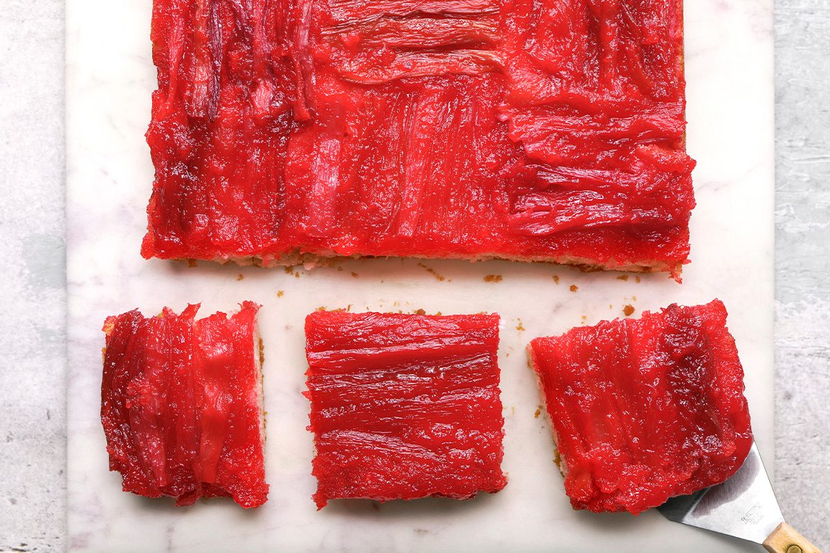 A rectangular slab of bright red dessert, likely made with watermelon, sits on a white surface. Two square pieces have been cut and separated from the main piece, with a metal spatula under one slice.