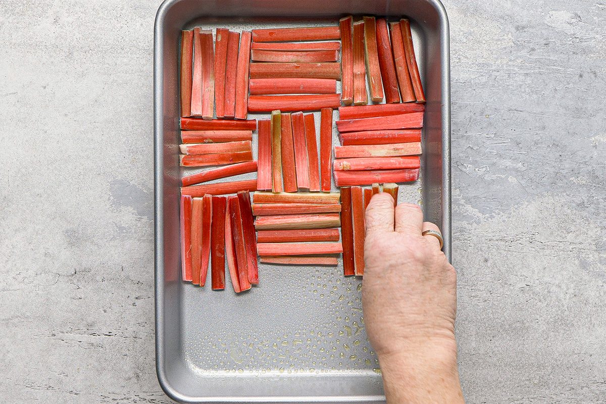 A hand arranges pieces of raw rhubarb in a neat, geometric pattern inside a rectangular metal baking pan on a light gray surface.