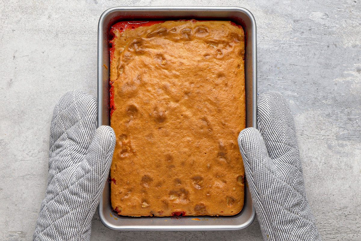 A person wearing gray oven mitts holds a rectangular baking pan containing a baked dessert with a golden-brown, textured top. The pan is set on a light gray surface.