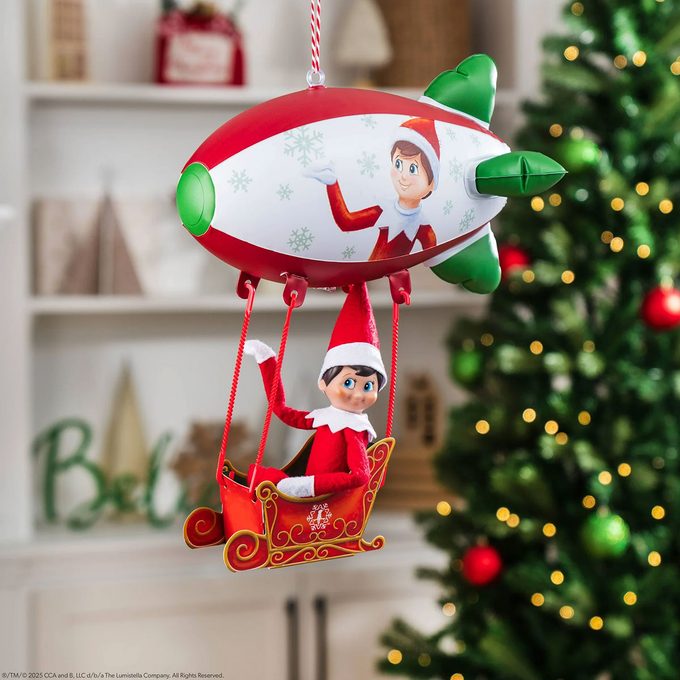 A Christmas tree ornament featuring an Elf on the Shelf sitting in a red sleigh, which is suspended from a rocket-shaped airship decorated with snowflakes, with a decorated Christmas tree in the background.