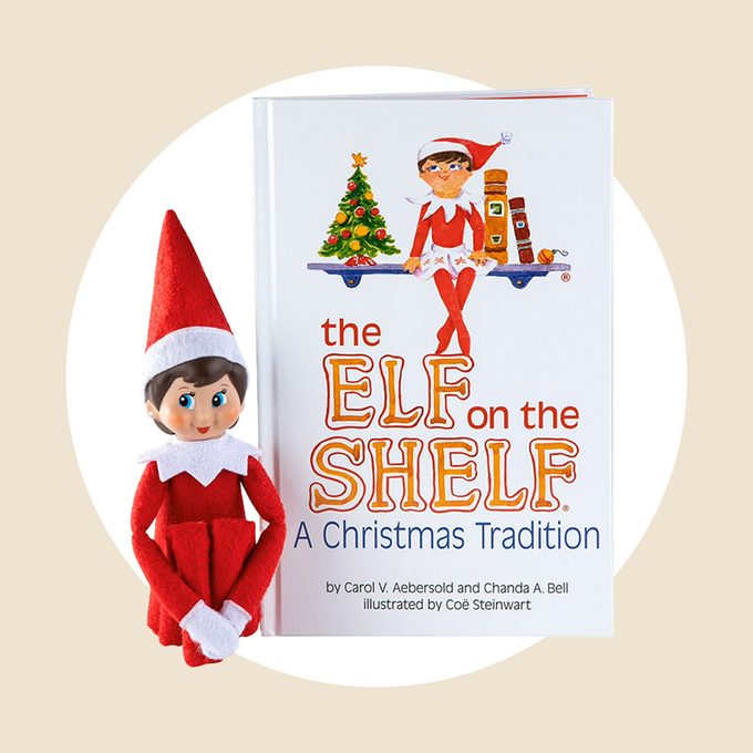 A plush Elf on the Shelf doll sits next to a book titled "The Elf on the Shelf: A Christmas Tradition," set against a light beige circular background.