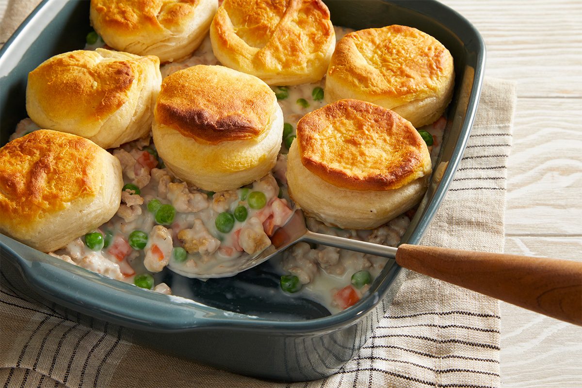 A baking dish filled with creamy chicken and vegetable pot pie topped with golden-brown biscuit rounds, with a serving spoon scooping out a portion. The dish sits on a striped cloth.