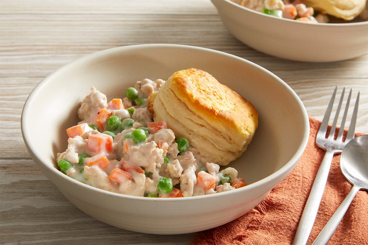 A bowl of creamy chicken and vegetable stew with peas and carrots, topped with a golden biscuit. A fork and spoon rest nearby on an orange napkin.