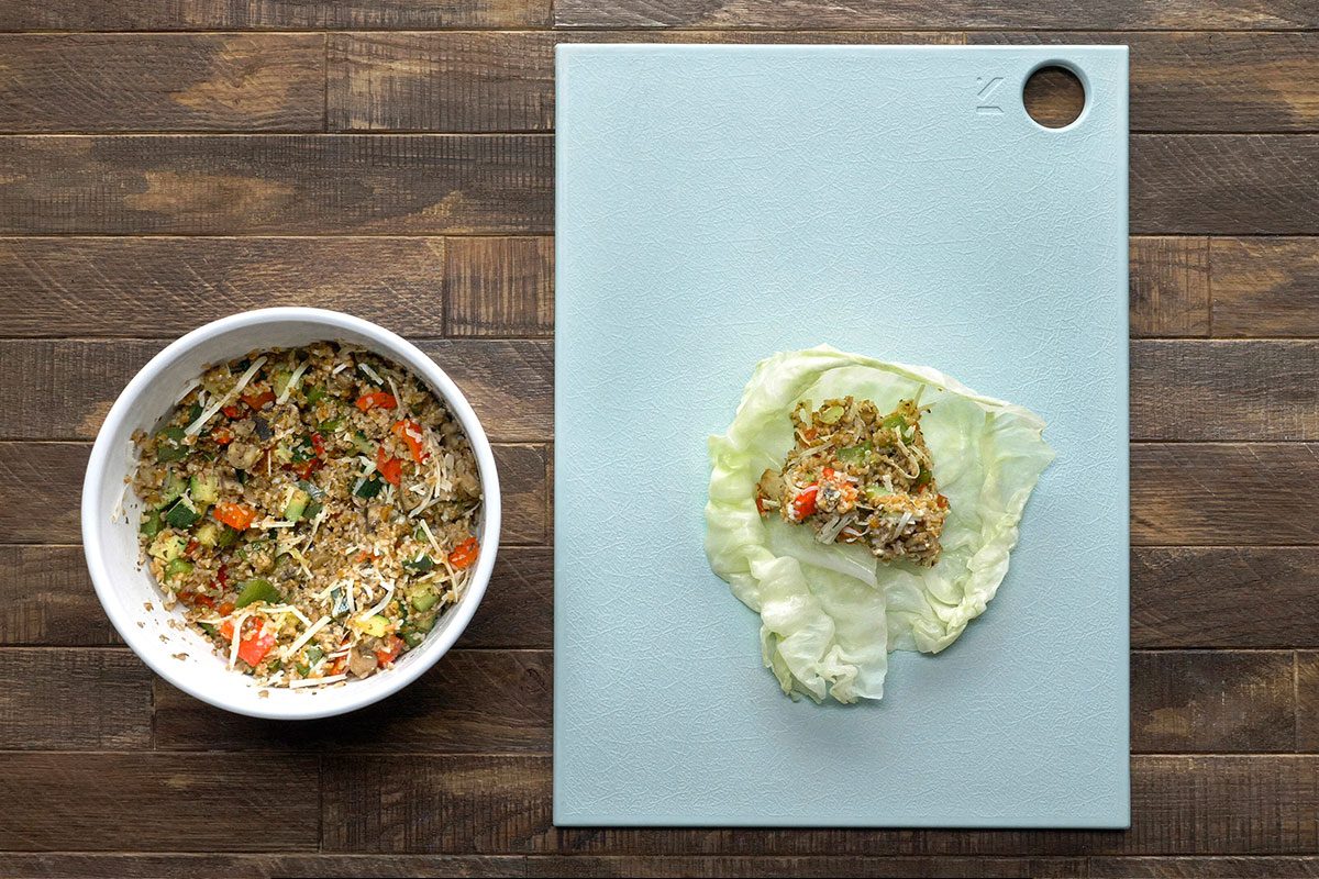 A bowl of vegetable and noodle mixture sits on a wooden table next to a white cutting board, where a portion of the mixture is placed on a lettuce leaf, ready to be rolled.