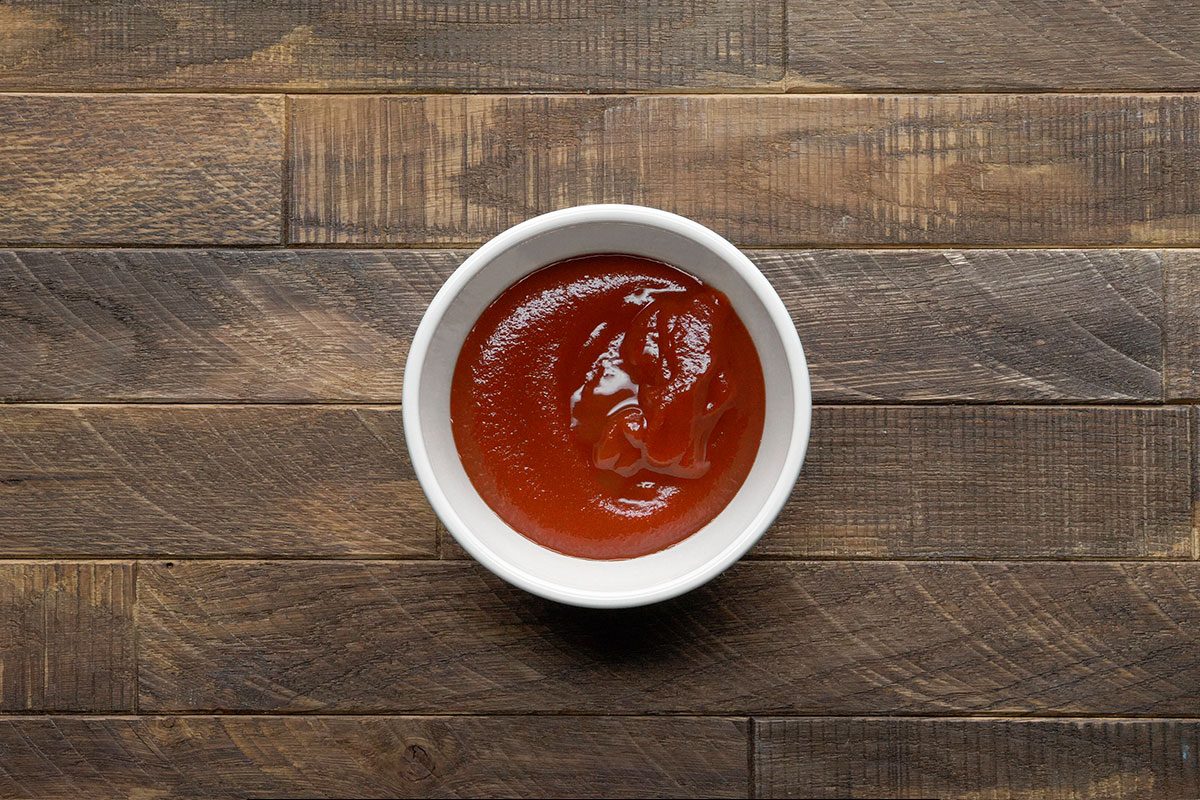 A small white bowl filled with tomato ketchup sits on a rustic wooden surface.