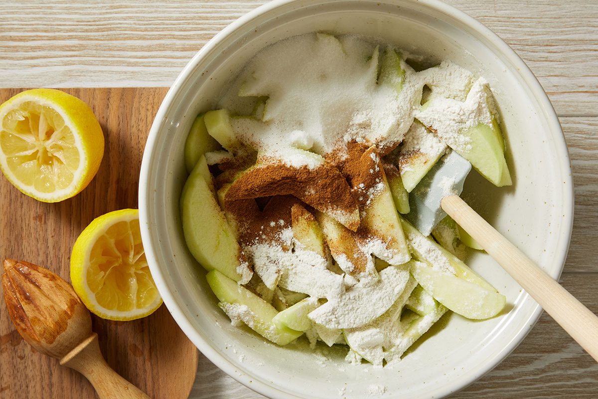 A mixing bowl filled with sliced green apples, sugar, flour, and cinnamon, next to a cutting board with halved lemons and a wooden citrus reamer.