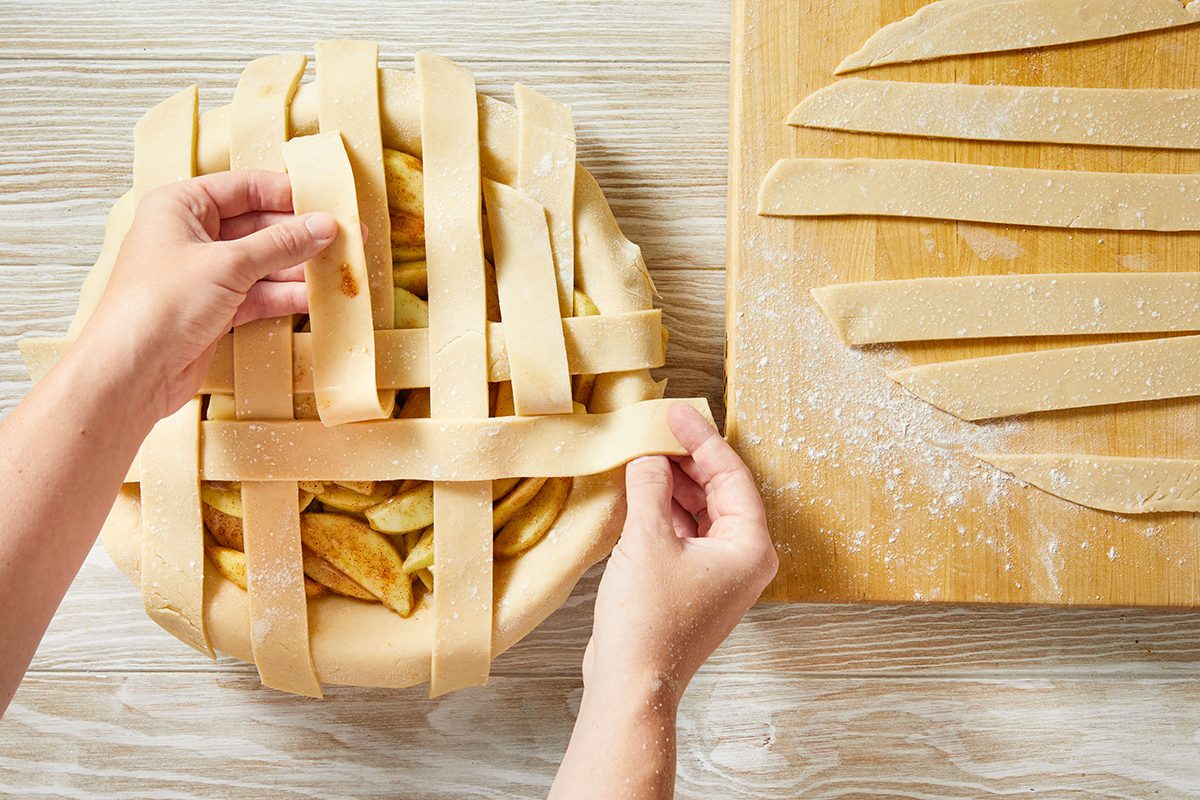 Hands weaving strips of dough into a lattice pattern on top of an unbaked pie; more dough strips are on a floured wooden board beside the pie.