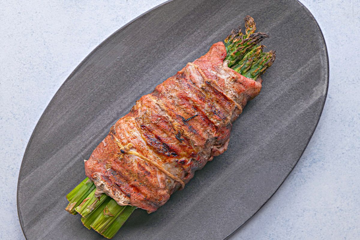 overhead shot of Asparagus Stuffed Pork Tenderloin; grilled asparagus spears wrapped in bacon, served on a dark oval plate on a light gray surface
