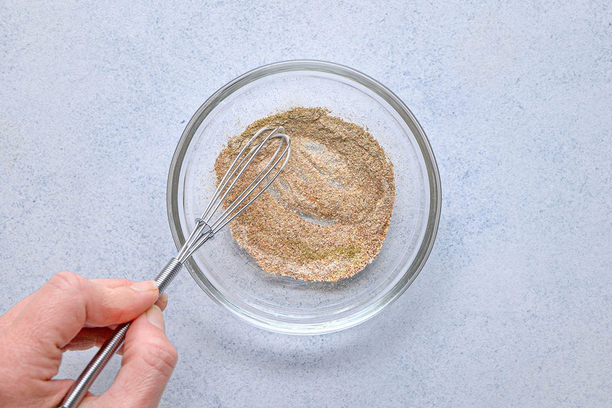 overhead shot of A hand holds a small whisk, mixing a dry seasoning blend in a clear glass bowl on a light gray surface
