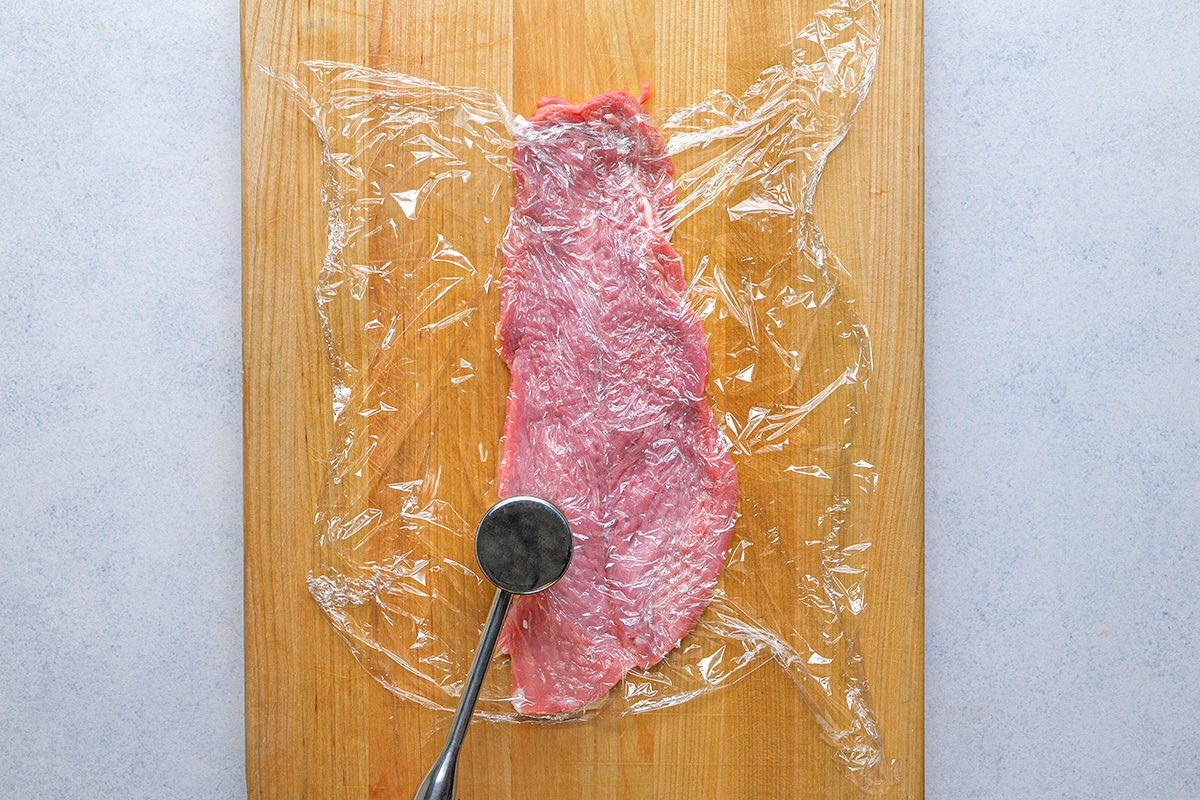 overhead shot of A raw piece of meat on a cutting board is covered with plastic wrap; A meat tenderizer mallet is pressing down on the meat