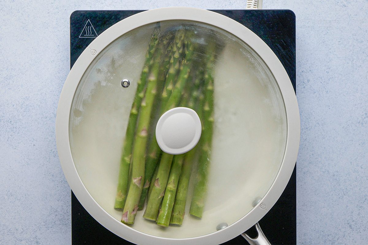 overhead shot of Spears of asparagus are being steamed in a pan covered with a glass lid on an electric stovetop
