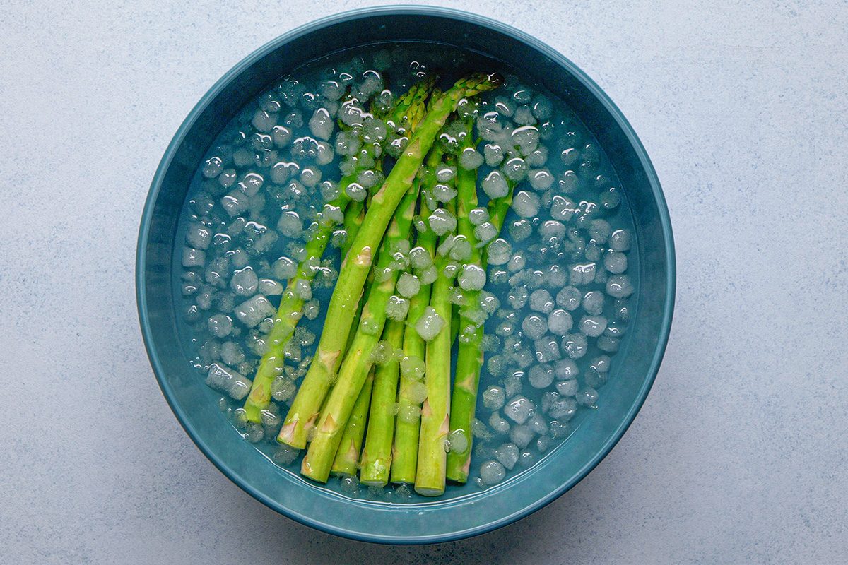 overhead shot of Bright green asparagus spears are submerged in a blue bowl filled with ice water and ice cubes, The background is a light, textured surface