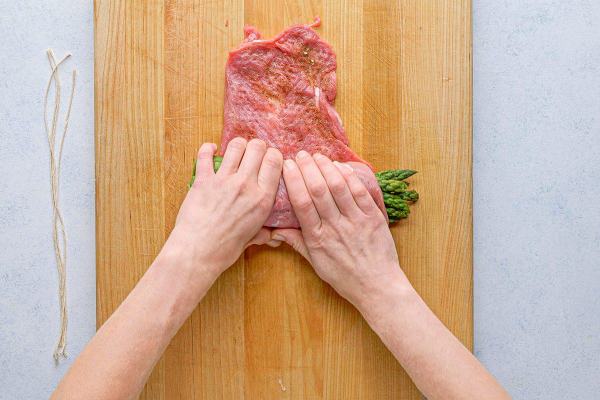 overhead shot of Hands are rolling a slice of raw meat around asparagus spears on a wooden cutting board, with two pieces of kitchen twine placed to the side