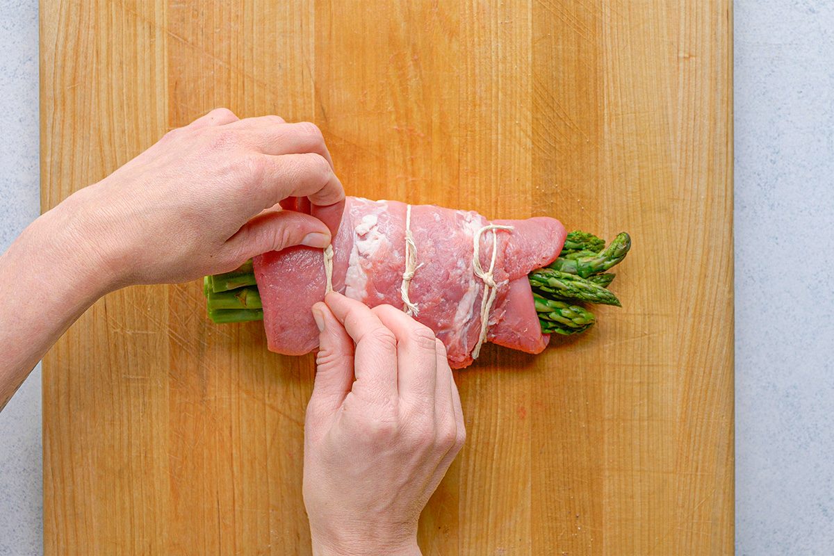 overhead shot of Hands tying kitchen twine around a bundle of asparagus wrapped in slices of raw meat on a wooden cutting board