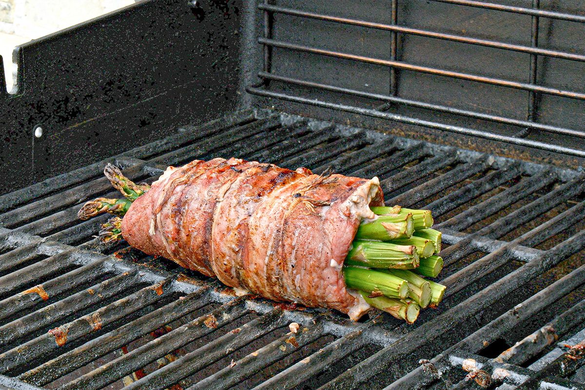 3/4th shot of A bundle of asparagus wrapped in strips of grilled steak is cooking on an outdoor gas grill; The steak is browned and the asparagus tips poke out from both ends