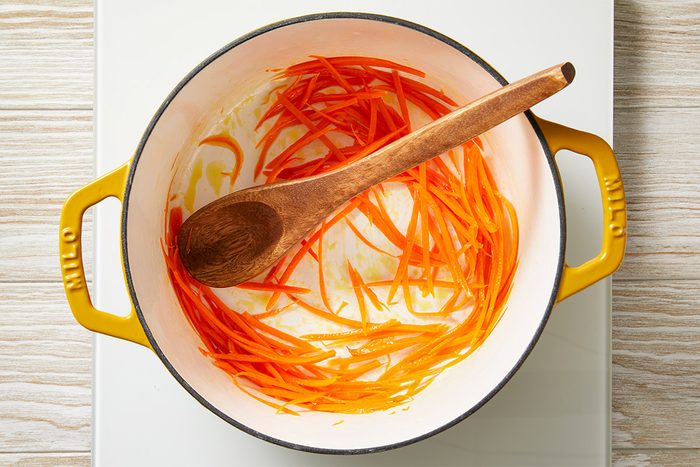 Overhead shot of a yellow-handled pot on a white stovetop, with thin carrot strips being sautéed and stirred with a wooden spoon;