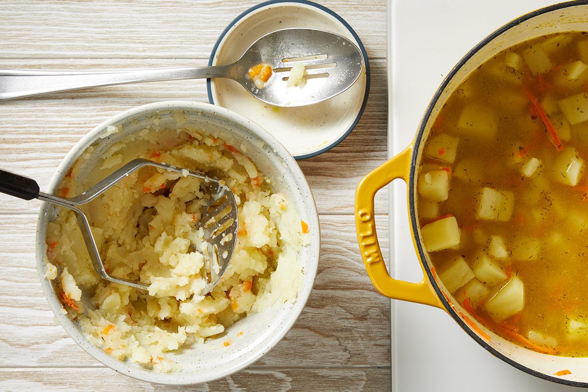 Overhead shot of a bowl of mashed potatoes with a masher, a plate with a fork, and a yellow pot of vegetable soup with visible potato chunks, all set on a light wooden surface;