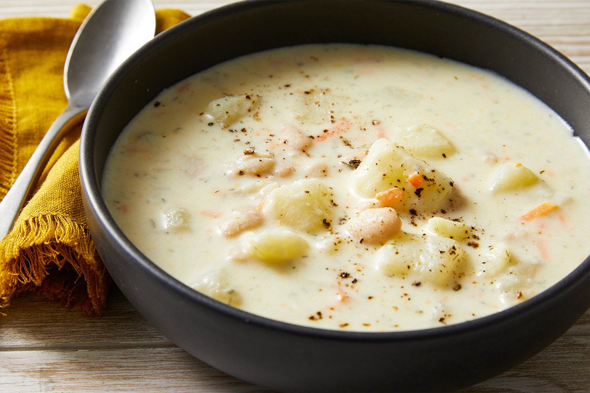 Close-up shot of a black bowl of Potato Soup with Beans topped with black pepper, set on a wooden surface beside a yellow napkin and a spoon;