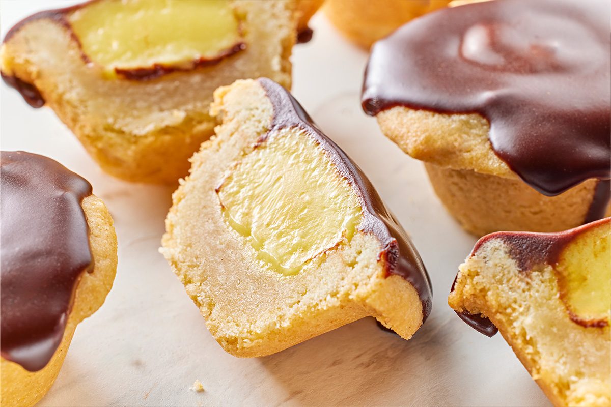 Closeup shot of a halved Boston Cream Pie Cookie, showing the soft golden cookie shell, smooth custard center, and glossy chocolate topping, with whole cookies surrounding it;