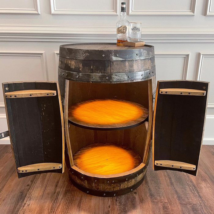 A wooden barrel converted into a storage cabinet with two shelves and open doors. Two bottles and two glasses sit on top. The cabinet sits on a wooden floor against a paneled wall.