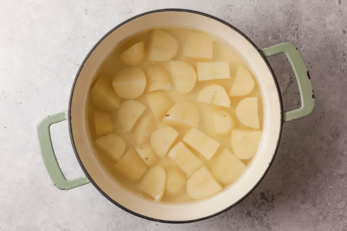 Overhead shot of a pot with green handles filled with peeled, chopped potatoes submerged in water, set on a light gray textured surface, ready for boiling;