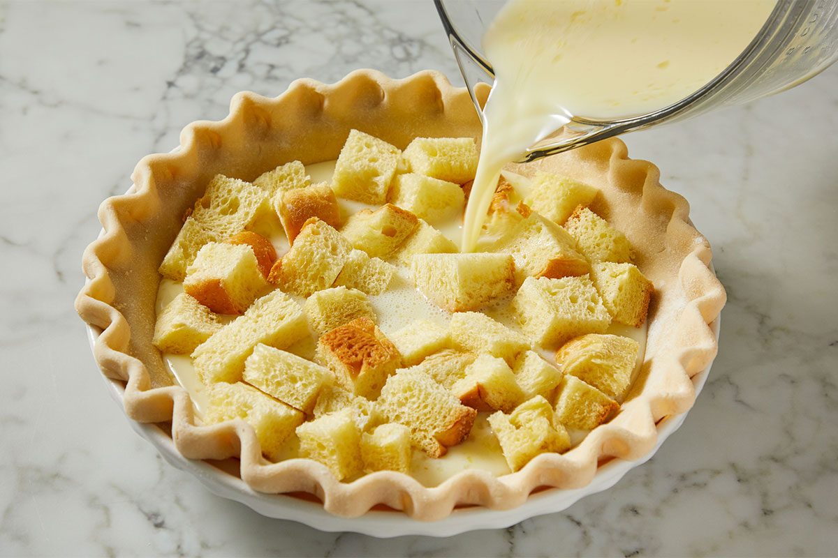 A pie crust filled with bread cubes sits on a marble surface as a creamy liquid is being poured over the bread from a glass measuring cup.